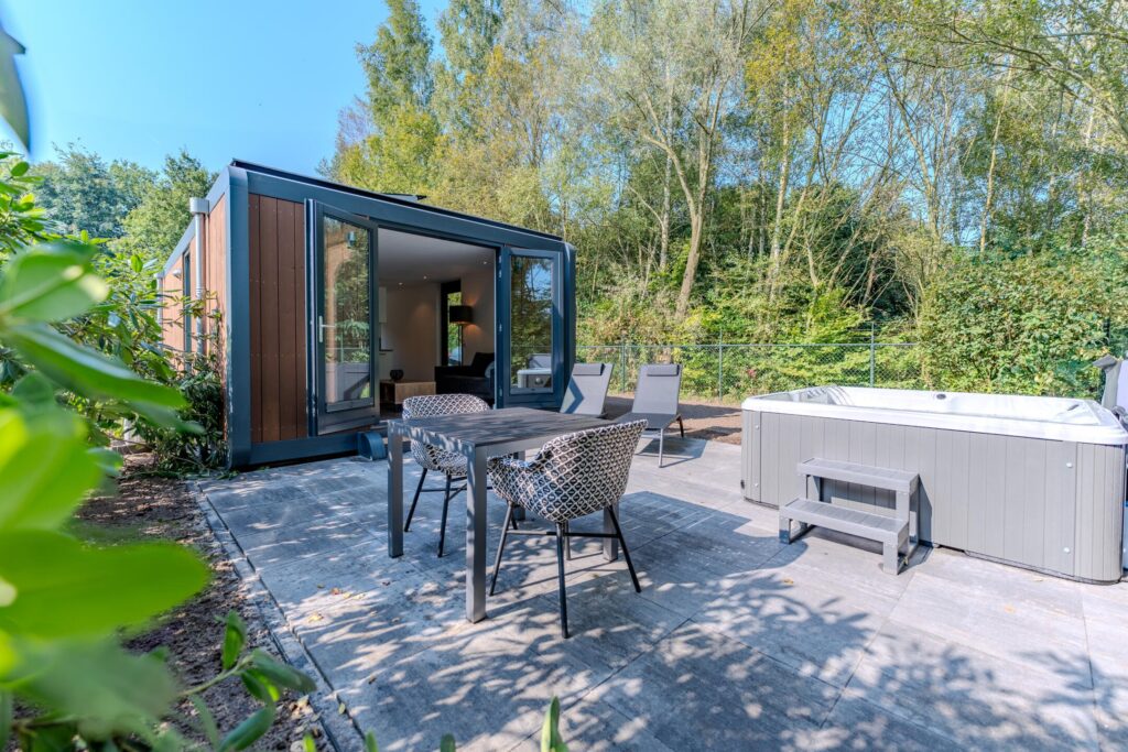 Moderne vakantiewoning met open glazen schuifdeur, buitenterras met tuinmeubilair en een jacuzzi, omgeven door natuur.