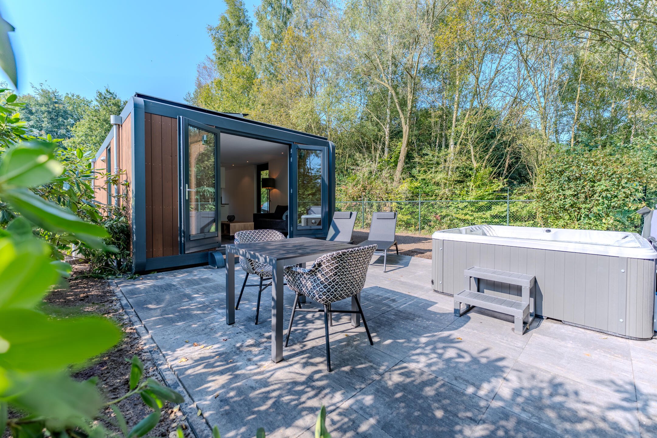 Moderne vakantiewoning met open glazen schuifdeur, buitenterras met tuinmeubilair en een jacuzzi, omgeven door natuur.